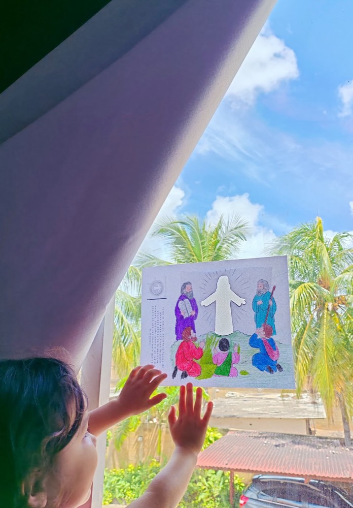 Una niña pequeña pega en la ventana su dibujo coloreado de la Transfiguración para que le pase la luz